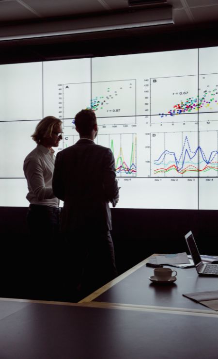 Two professionals analyzing data on large screens in a modern conference room, with graphs illustrating cybersecurity metrics and trends, reflecting the importance of CMMC compliance for defense contractors.